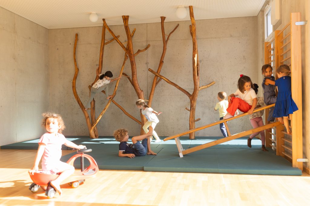 Neun Kinder klettern und spielen im Gruppenraum der kinderBlüte: auf Holzkonstrukten, einer Kletterwand, einer Bank und einem Plastikfahrzeug.