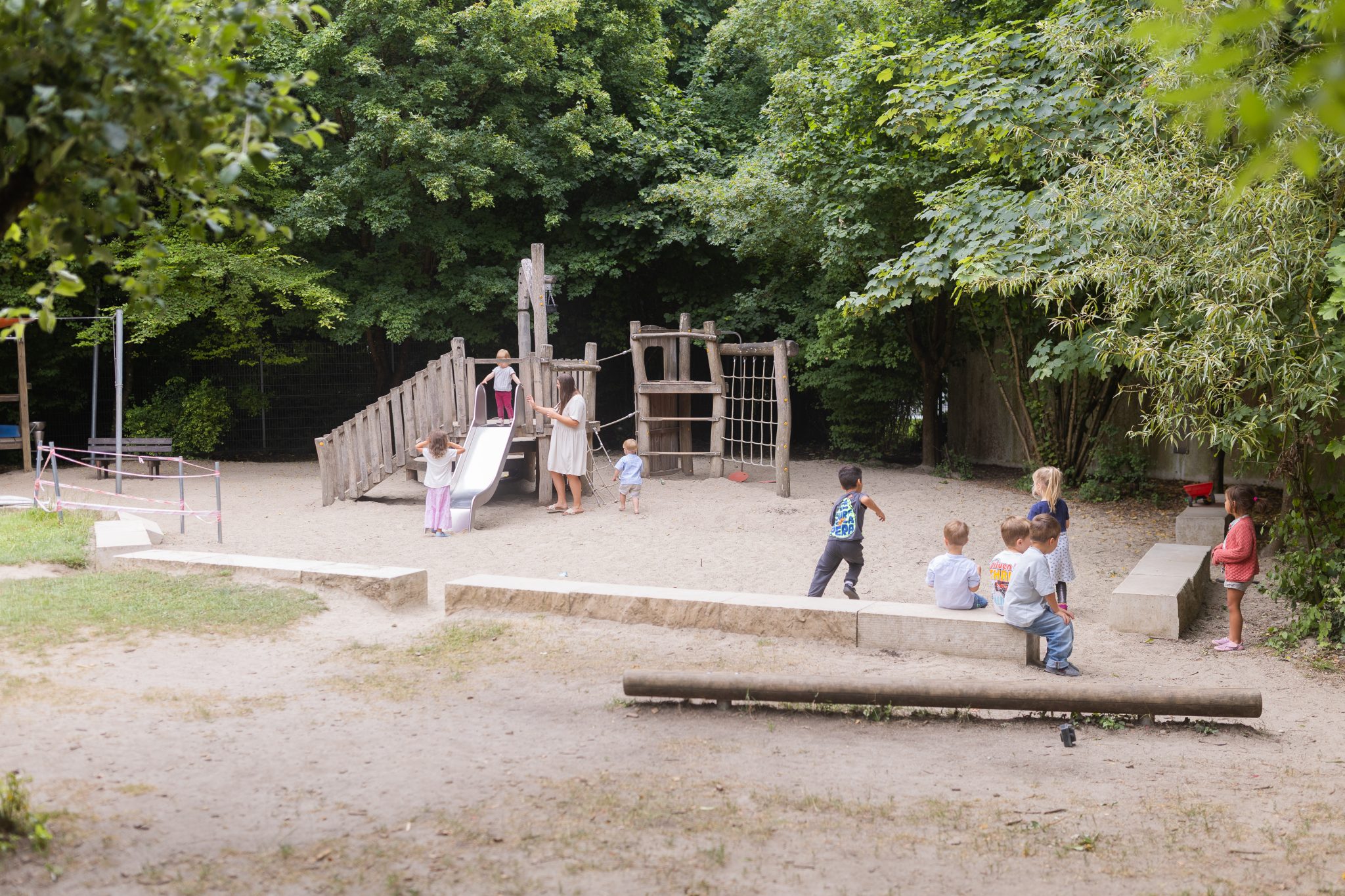 Mehrere Kinder spielen und bewegen sich auf einem sandigen Außengelände mit Holzspielgeräten und Rutsche.
