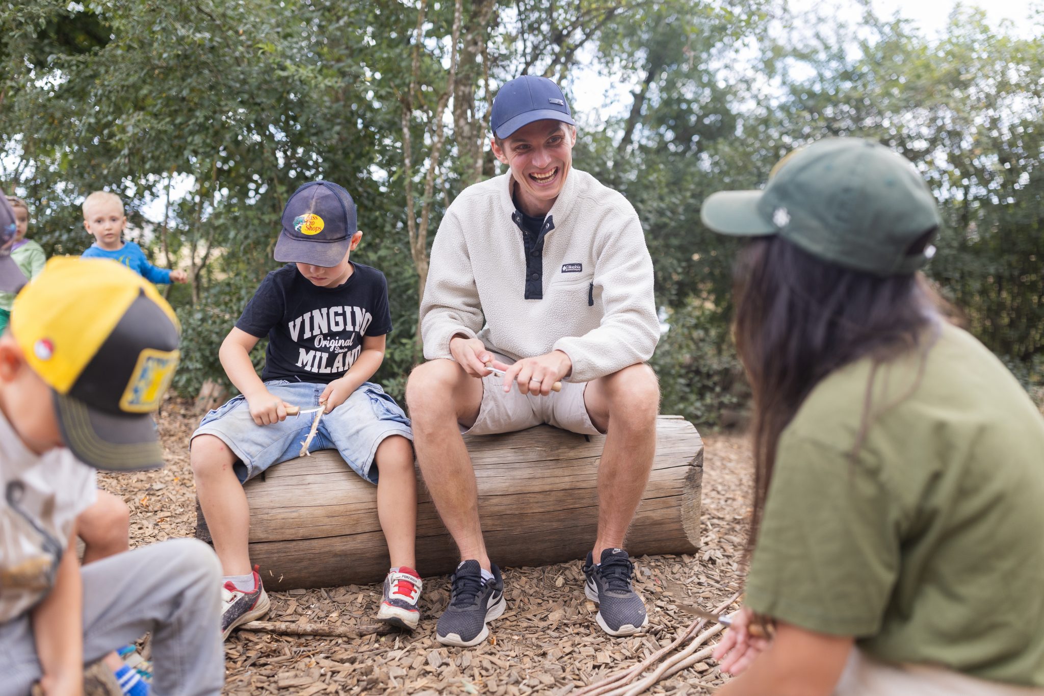 Pädagogische Fachkräfte sitzen mit Kindern auf Baumstämmen im Wald und beschäftigen sich gemeinsam mit Naturmaterialien.