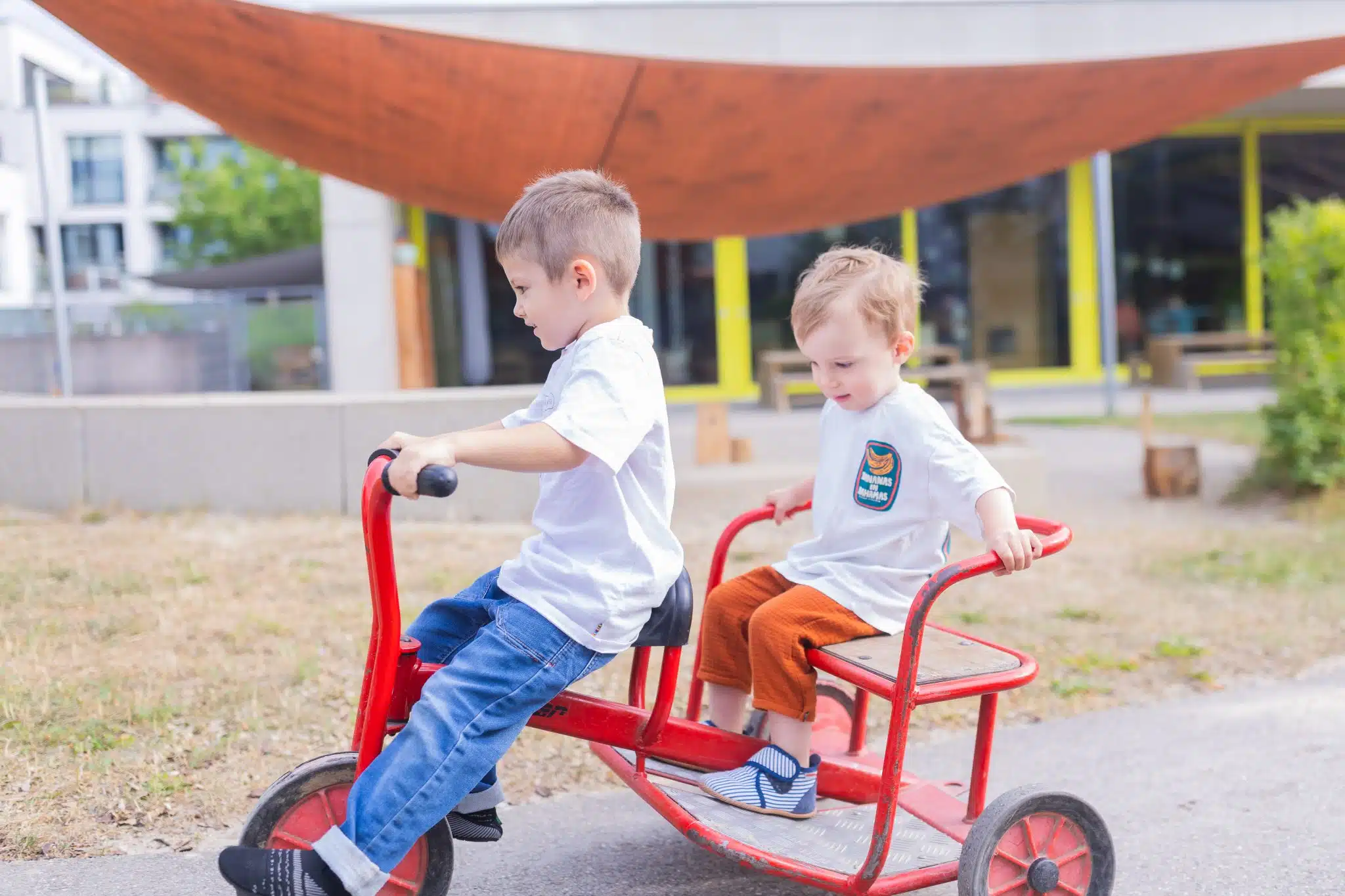 Zwei Kinder fahren auf einem roten Kinderfahrzeug im Garten.
