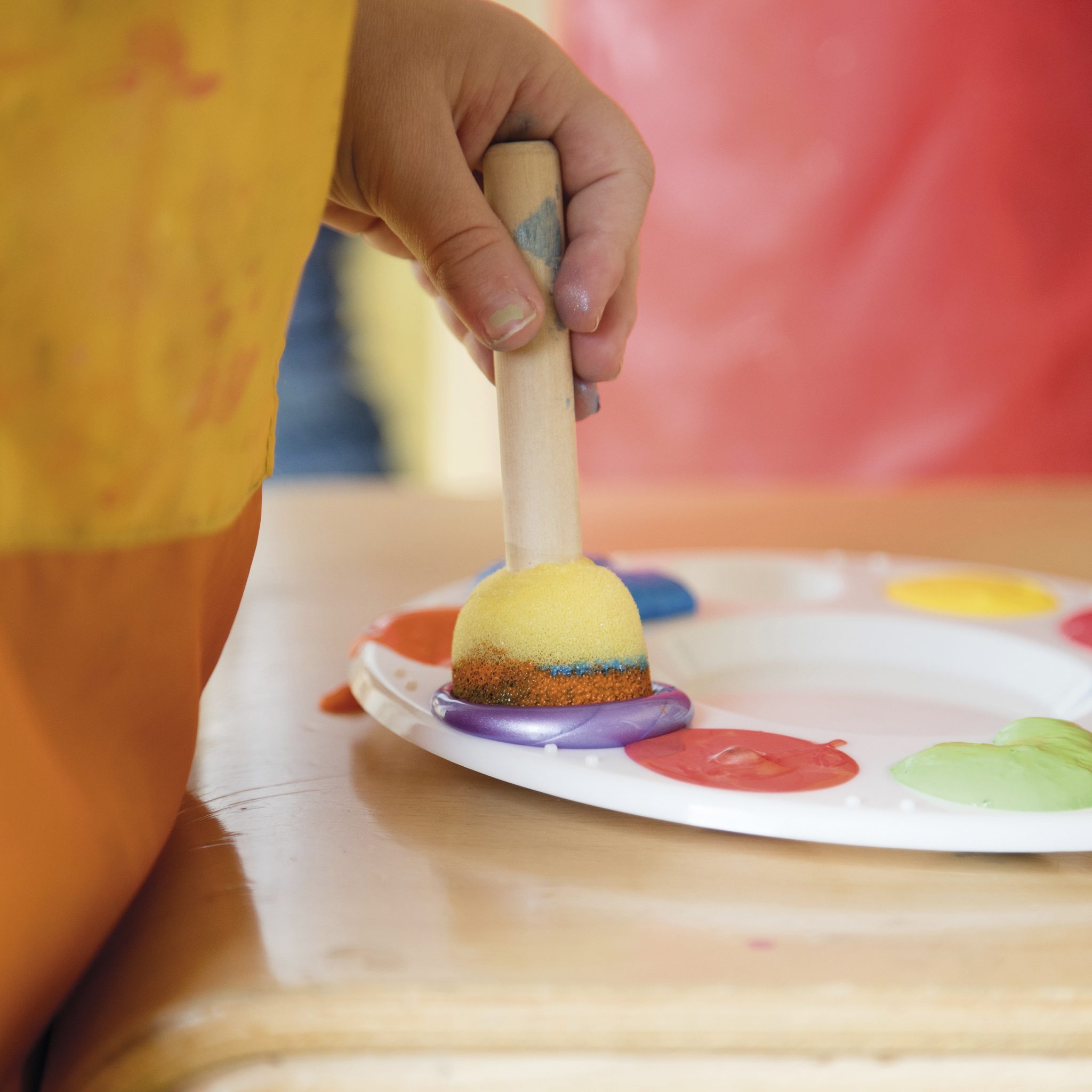 Kinderhand taucht einen Schwammstempel in bunte Farben auf einer Malpalette – Profilbild der Kita kinderHaus.