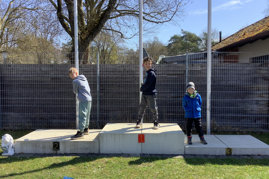 Drei Kinder auf einem Siegerpodest im Garten oder Schulhof bei sonnigem Wetter, spielen und lachen