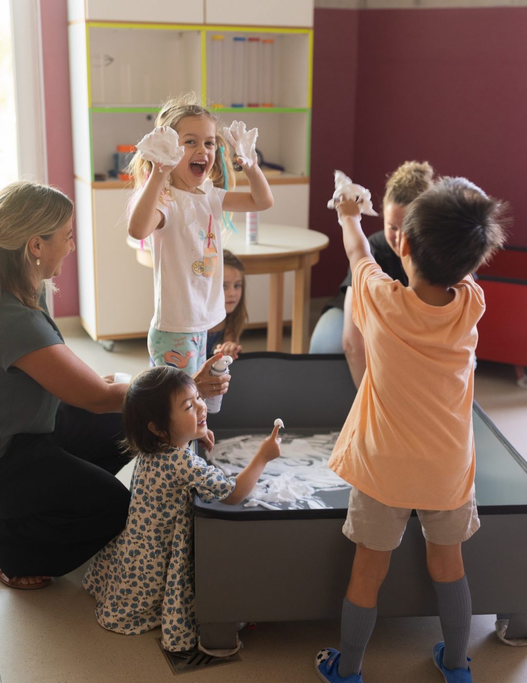 Kinder unterschiedlichen Alters spielen und experimentieren gemeinsam an einem Spieltisch im Innenraum einer Kindertagesstätte in Ingolstadt, begleitet von Erzieherinnen.