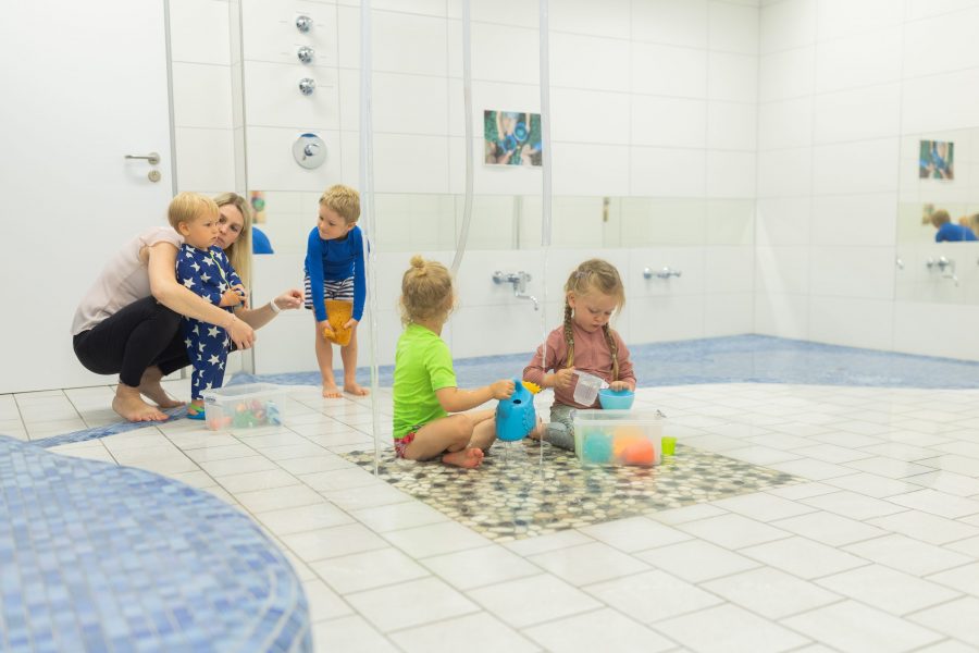 Mehrere Kinder sitzen und spielen mit Wasser und Behältern auf einem gefliesten Boden, während eine erwachsene Person sie begleitet.