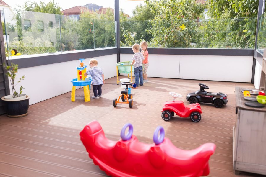Kinder spielen auf einer sonnigen Außenterrasse einer Kindertagesstätte der bürgerhilfe ingolstadt KiTa GmbH mit Fahrzeugen und Wasserspieltischen