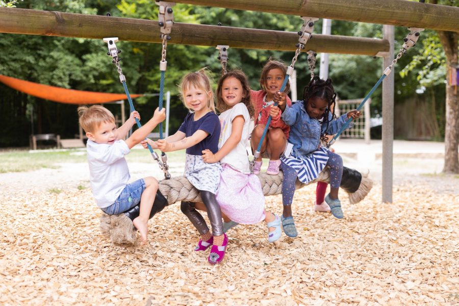 Mehrere Kinder sitzen nebeneinander auf einer Seilschaukel im Außenbereich und halten sich an Seilen fest.