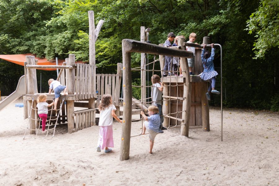 Mehrere Kinder klettern und spielen auf einem Holzspielgerät mit Netzen und Plattformen im Freien.