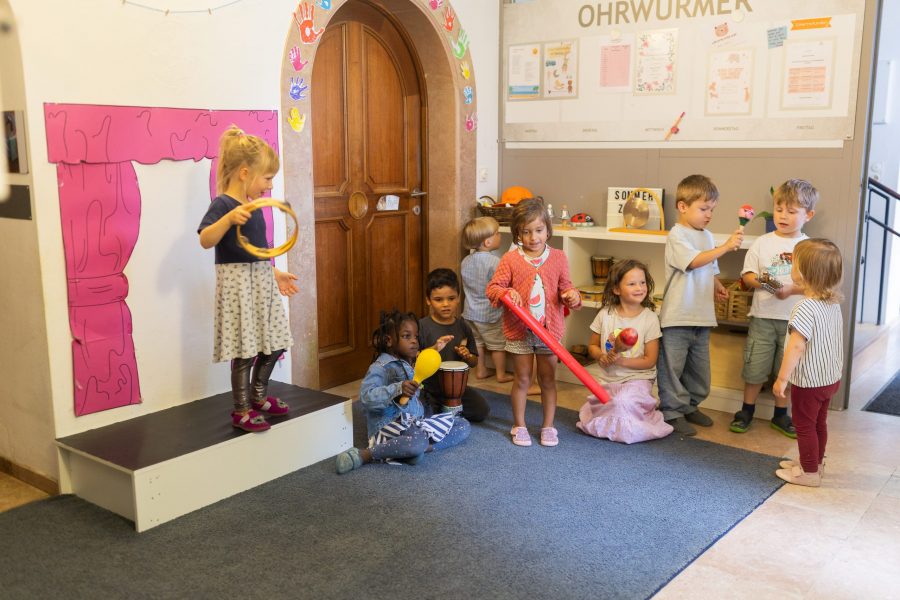 Mehrere Kinder stehen und sitzen in einem Raum und halten unterschiedliche Musikinstrumente in den Händen.