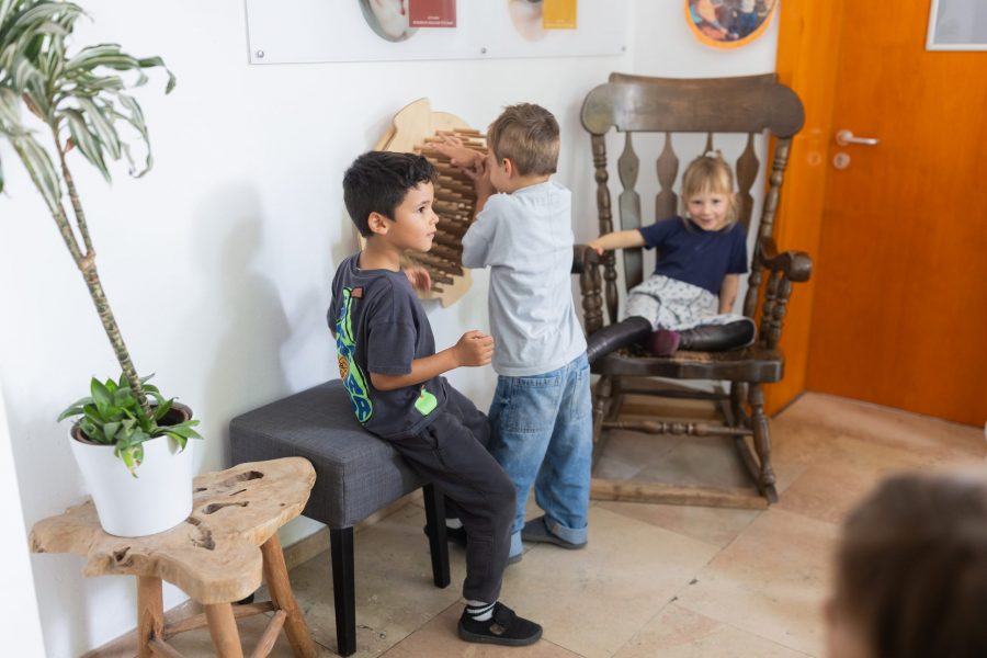 Drei Kinder befinden sich in einem Raum, eines sitzt auf einem Stuhl, zwei weitere spielen an einem hölzernen Wandelement.