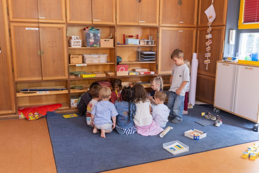 Mehrere Kinder sitzen auf einem Teppich vor einem Regal und spielen gemeinsam mit Materialien, ein Kind steht daneben.
