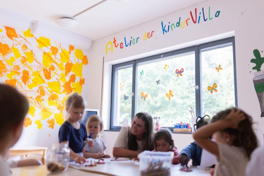 Mehrere Kinder und eine erwachsene Person sitzen an einem Tisch und beschäftigen sich mit kreativen Materialien in einem hellen Raum.