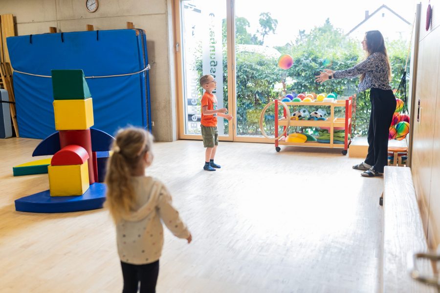 Kinder spielen gemeinsam mit einer Erzieherin ein Ballspiel im Bewegungsraum einer Kindertagesstätte der bürgerhilfe ingolstadt KiTa GmbH