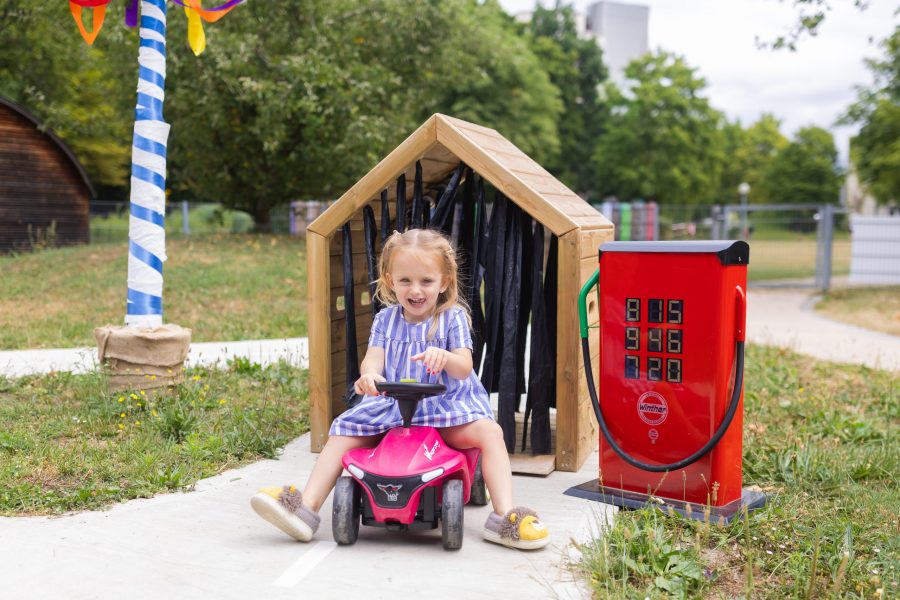 Ein Kind fährt mit einem Rutschfahrzeug im Außenbereich einer Kindertagesstätte in Ingolstadt und spielt an einer kindgerechten Spieltankstelle.