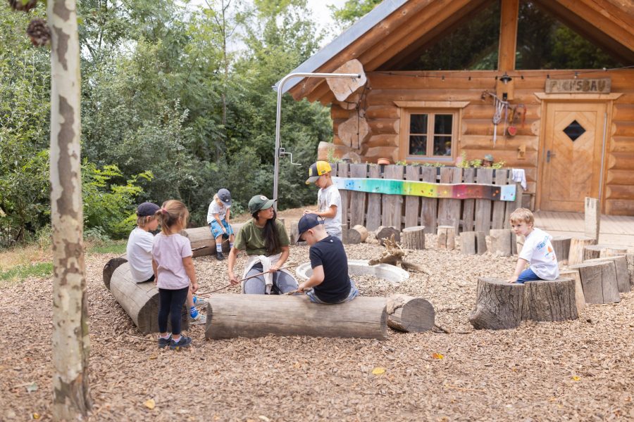 Kinder sitzen und spielen gemeinsam mit einer pädagogischen Fachkraft im naturnahen Außengelände einer Kindertagesstätte in Ingolstadt.