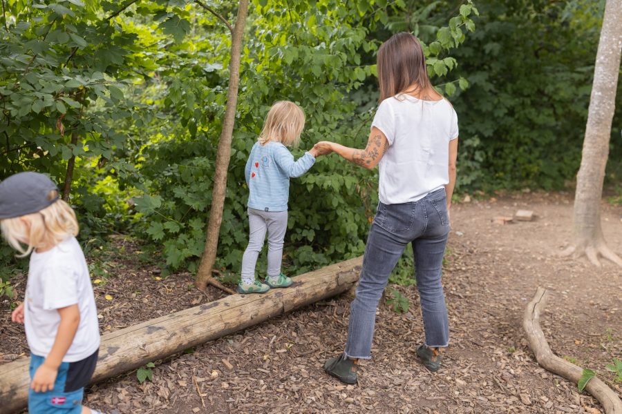 Eine pädagogische Fachkraft hält die Hand eines Kindes beim Balancieren auf einem Baumstamm im Wald einer Kindertagesstätte in Ingolstadt.