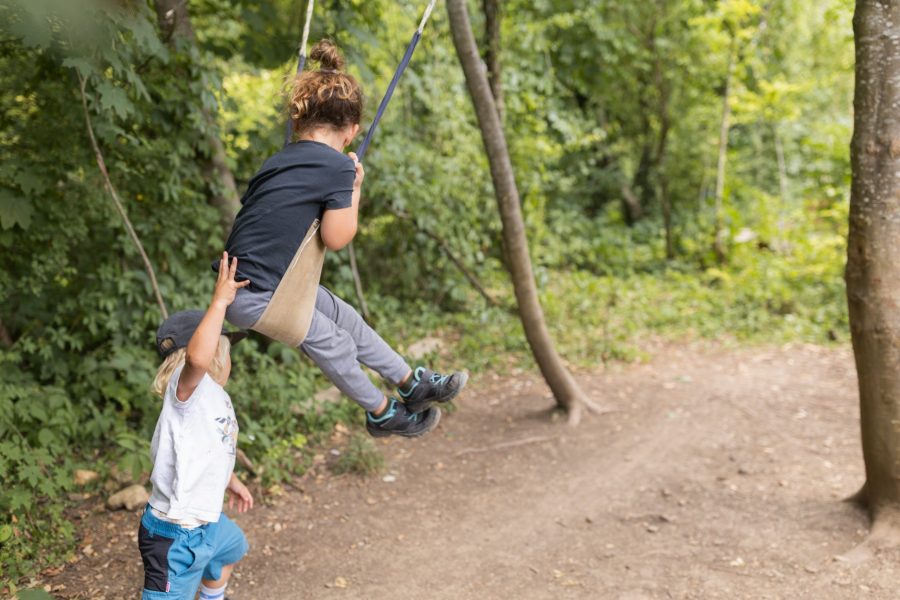 Zwei Kinder spielen im Wald, eines schaukelt an einem Seil, das andere gibt vorsichtig Schwung.