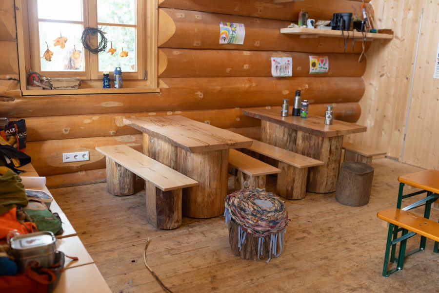Rustikaler Holzraum im KinderGrün mit Tischen, Bänken und Sitzhockern aus Baumstämmen.