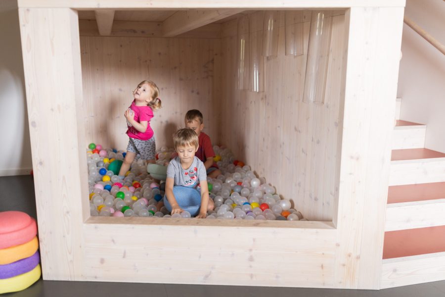 Drei Kinder spielen in einem hölzernen Bällebad mit bunten Kunststoffbällen.