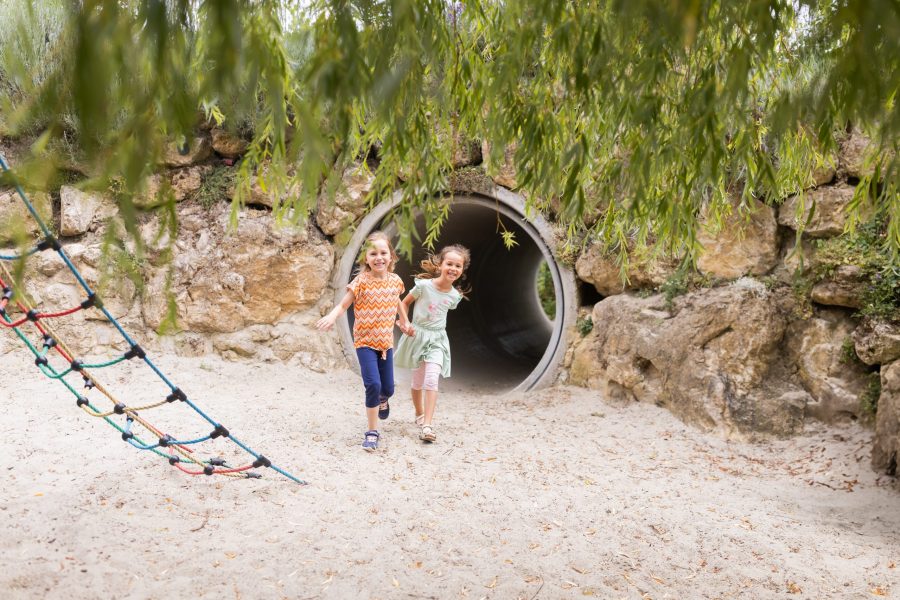 Zwei Kinder laufen aus einem Tunnel auf einem sandigen Spielplatz mit Naturmaterialien.