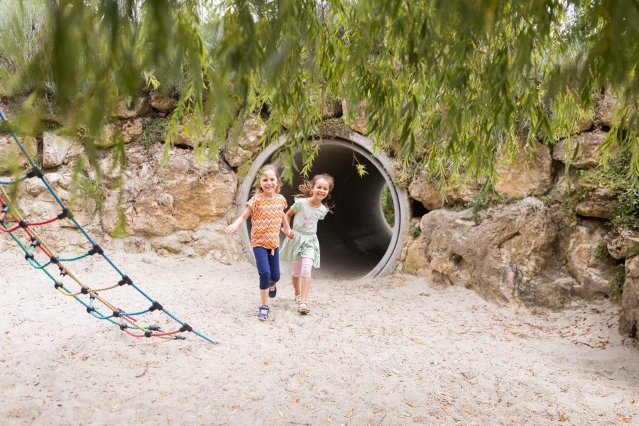 Zwei Kinder laufen lachend aus einem Tunnel auf dem Sandspielplatz.