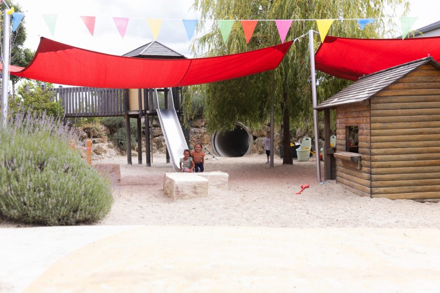 Kinder spielen auf einem Sandspielplatz mit Rutsche, Tunnel und Holzspielhaus unter Sonnensegeln.