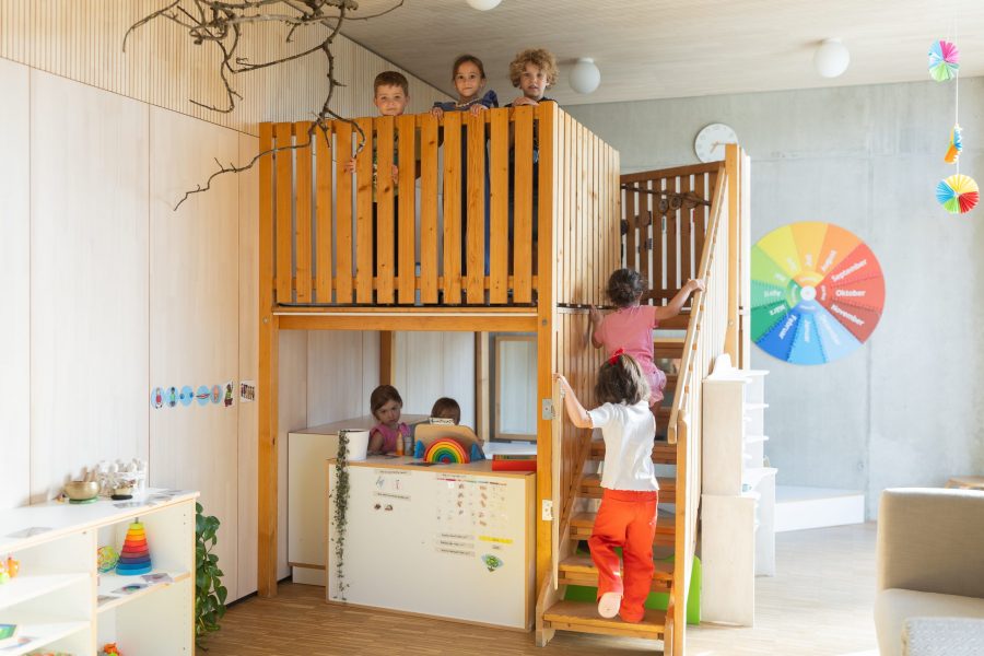 Sieben Kinder spielen gemeinsam auf, in und unter einem Holzkonstrukt in einem Gruppenraum der kinderBlüte und erkunden spielerisch verschiedene Ebenen.