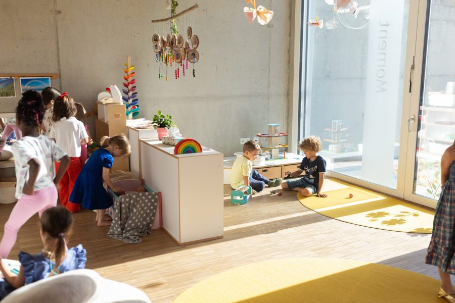 Acht Kinder spielen und beschäftigen sich frei im Gruppenraum der kinderBlüte, suchen Spielmaterial, spielen am Boden und lesen gemeinsam.