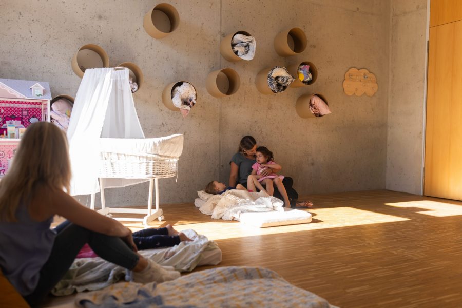 Drei Kinder und zwei Erzieher sitzen und liegen auf Matratzen im Gruppenraum der kinderBlüte und verbringen einen ruhigen, gemeinsamen Moment.