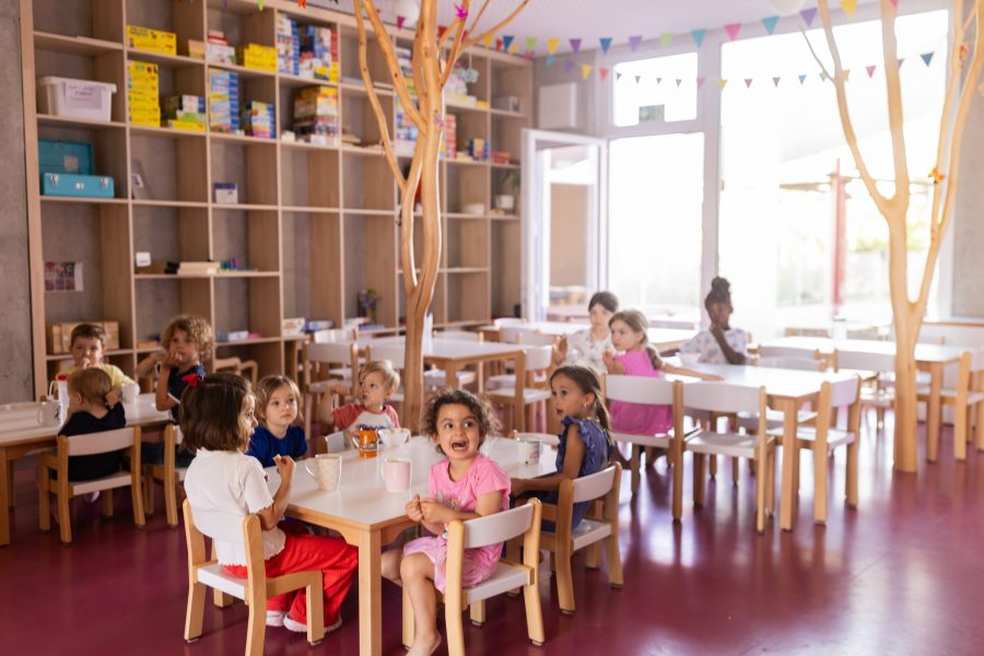 Zehn Kinder sitzen an Tischen in einem hellen Gruppenraum der kinderBlüte, trinken oder essen aus Schüsseln und Tassen, umgeben von Holzskulpturen und Spielregalen.