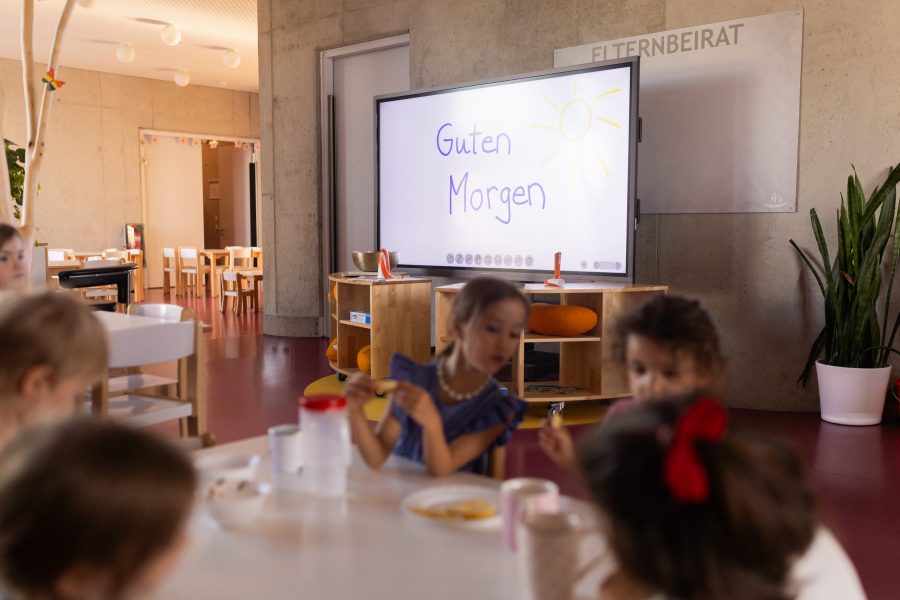 Fünf Kinder sitzen um einen Tisch und essen gemeinsam, im Hintergrund zeigt ein Fernseher „Guten Morgen“ in blauer Schrift mit einer gelben Sonne.