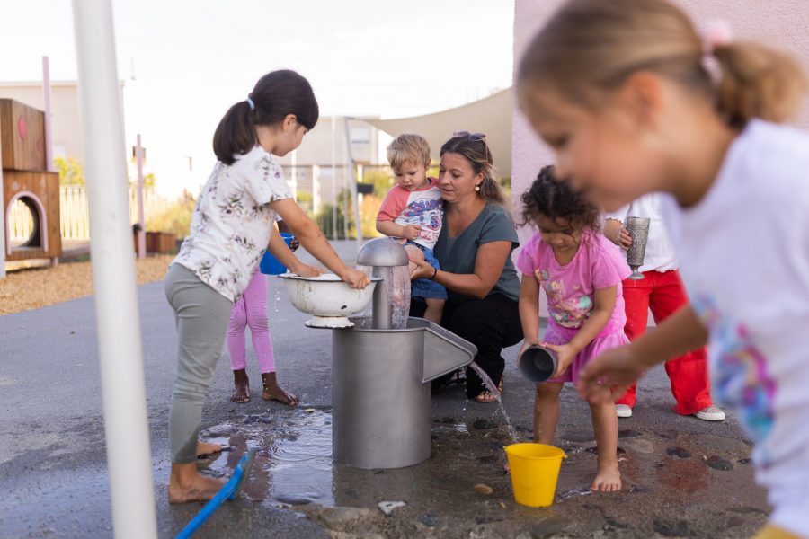 Kinder verschiedener Altersgruppen spielen gemeinsam an einer Wasserstation im Außenbereich einer Kindertagesstätte in Ingolstadt, begleitet von einer Erzieherin.
